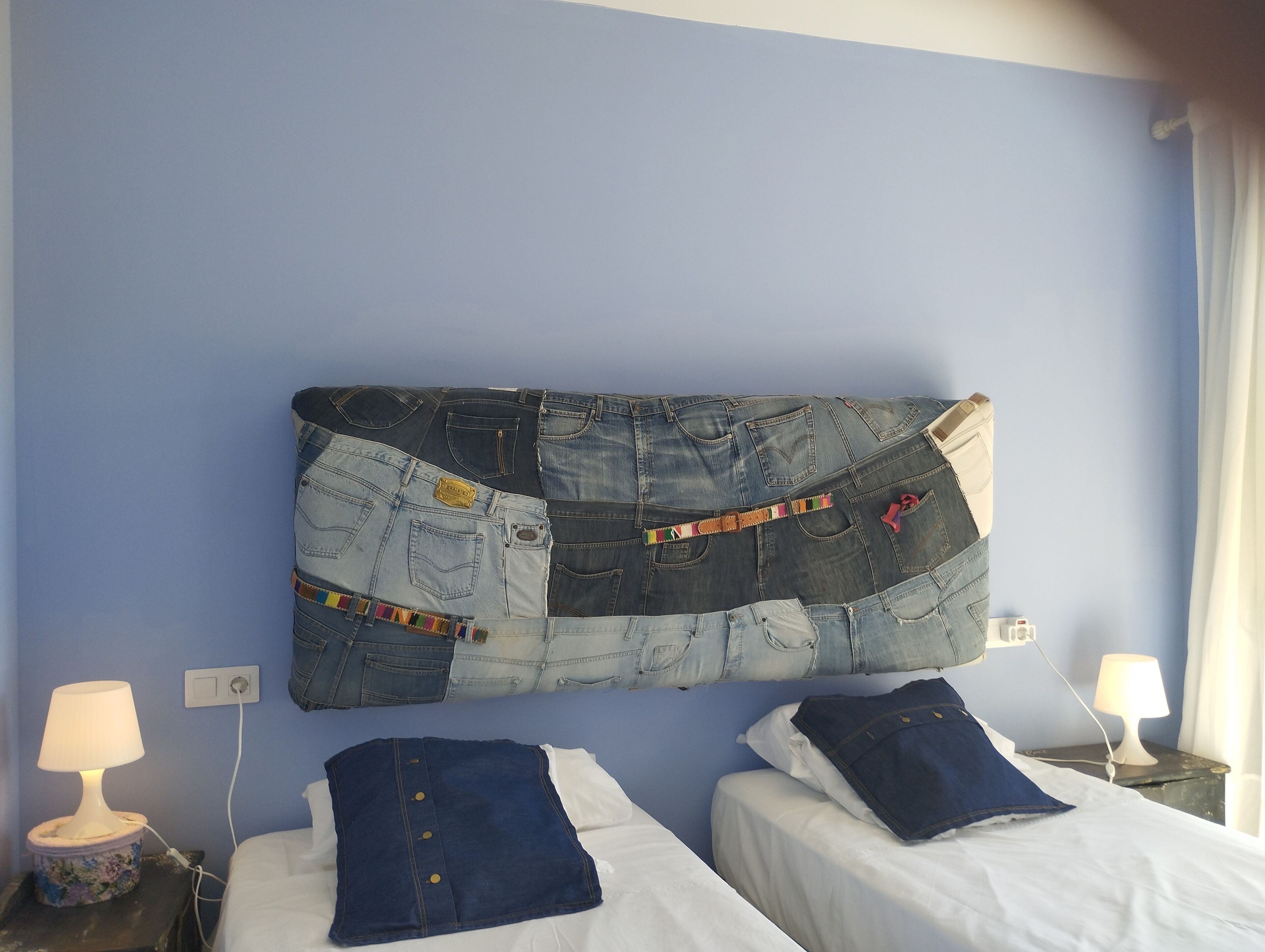 Een kamer met twee bedden, een wanddecoratie van denim en een nachtkastje met een lamp.