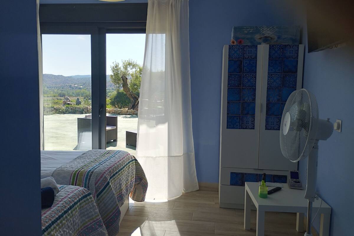 A bedroom with a bed, a desk, a chair, a fan, and a view of the outdoors.