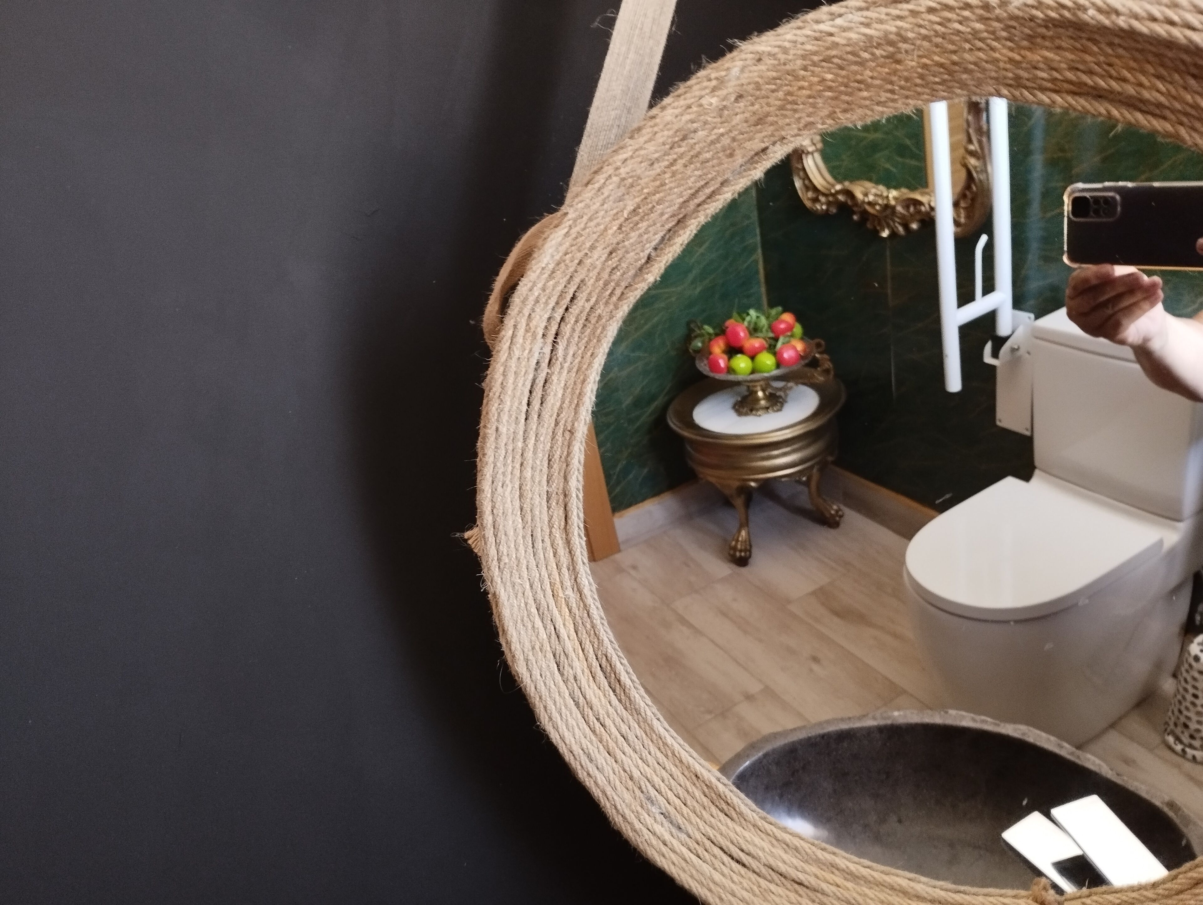A bathroom with a round mirror, a toilet, a small table with a fruit bowl, and a decorative rope on the wall.