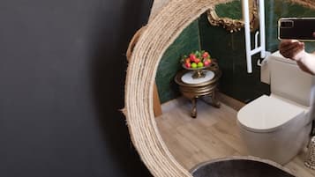 A bathroom with a round mirror, a toilet, a small table with a fruit bowl, and a decorative rope on the wall.