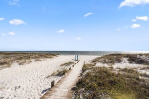 A sandy beach with a wooden boardwalk leading to a blue structure, likely a lifeguard station, under a clear sky.