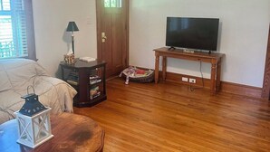 Ein Zimmer mit Deckenventilator, einem Fernseher auf einem Holzboden, einem Bett mit beiger Bettdecke, einem kleinen Tisch mit Laterne und einer Tür mit Fenster.
