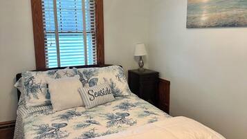 Un dormitorio con cama, ventana con persianas, mesita de noche con lámpara y un cuadro en la pared.