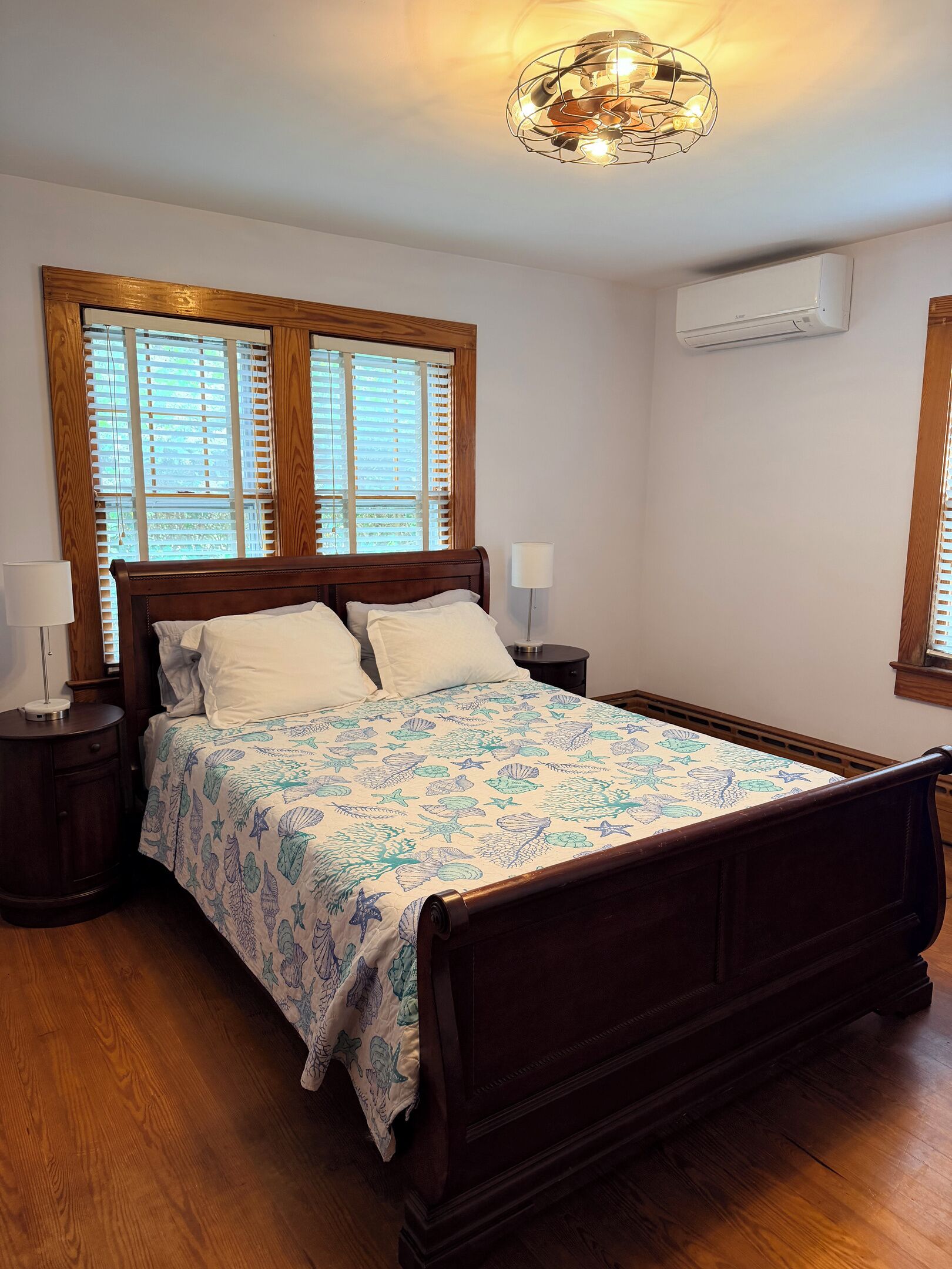 A bedroom with a wooden bed, a patterned bedspread, a nightstand with a lamp, and a ceiling light fixture.