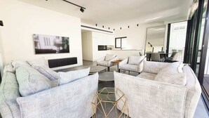 A modern living room with a flat-screen TV, grey upholstered sofas, a coffee table, and a hexagonal glass side table.