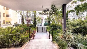 A covered outdoor pathway leading to a gated entrance, surrounded by lush greenery and residential buildings.