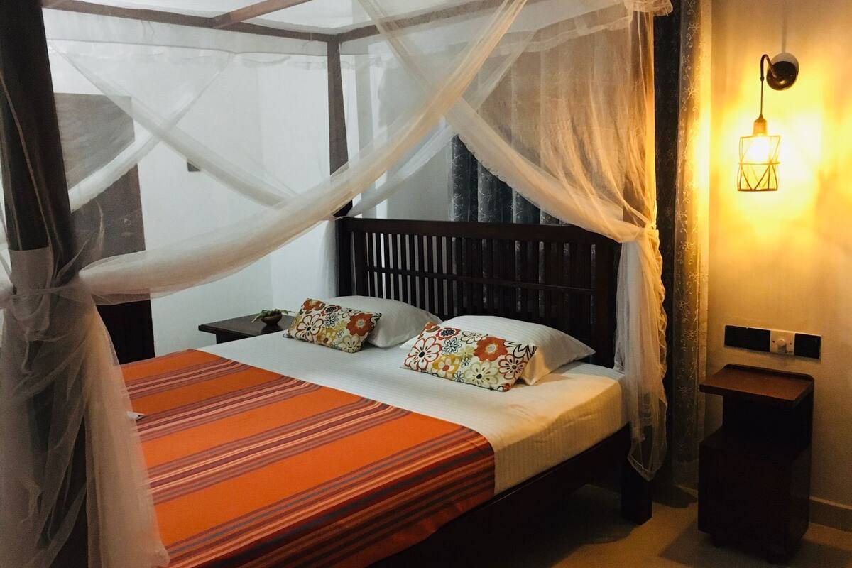 A bed with a canopy, a bedside table, a nightstand, a wall-mounted lamp, and a window with curtains.