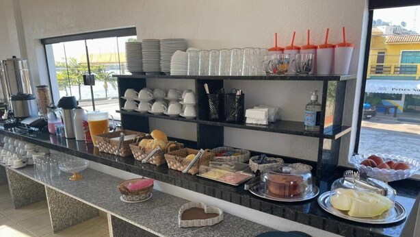 A buffet counter with various food items, including bread, pastries, and fruits. There are also several cups and plates stacked on shelves behind the counter. - Cananéia (Cananeia)