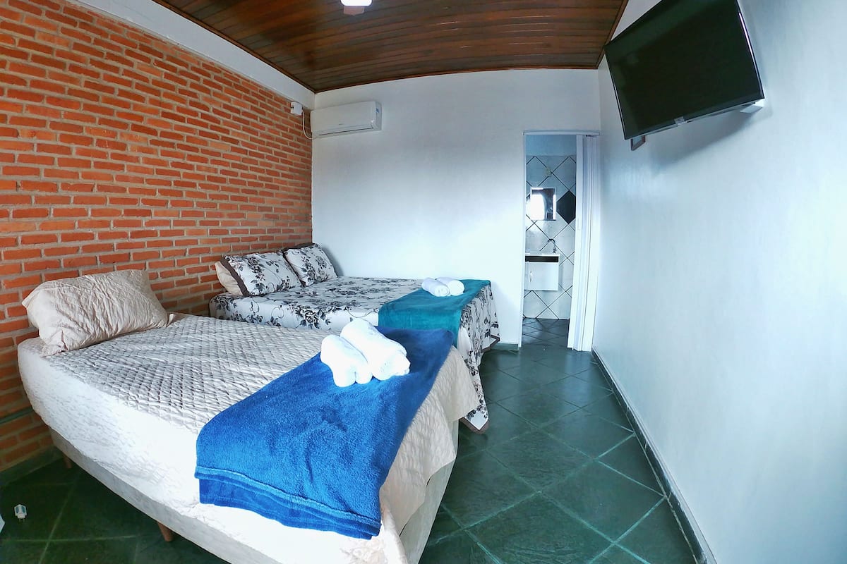 Quarto com duas camas, televisão fixada na parede e parede de tijolos.