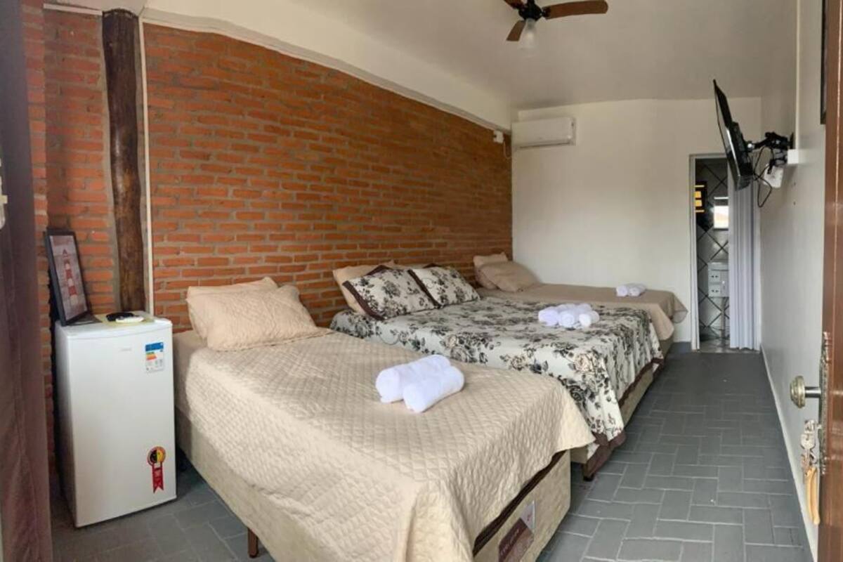 Quarto com duas camas, parede de tijolos, ventilador de teto e uma pequena geladeira.