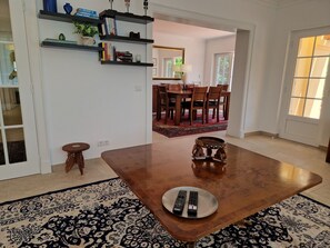 A dining area with a wooden table, two remote controls on a plate, a patterned rug, a small stool, and a bookshelf with books and decorative items.