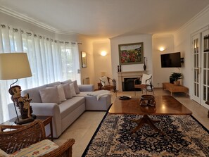 Ein Wohnzimmer mit einer Couch, Sesseln, einem Couchtisch, einem Kamin, einem Fernseher und einem großen Fenster mit Vorhängen.