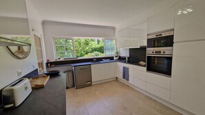 A modern kitchen with white cabinets, a built-in oven, a dishwasher, and a window overlooking greenery.