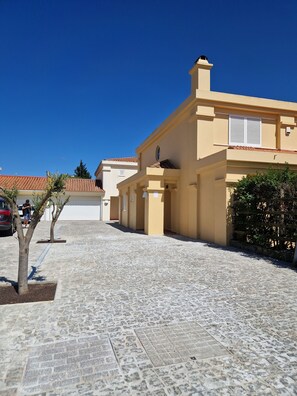 A paved street with buildings, a clear blue sky, and a few trees.