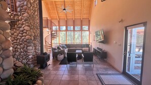 A spacious living area with a stone wall, a wooden ceiling, a fireplace, a sofa, a TV, and a staircase leading to an upper level.
