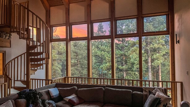A cozy living room with a wooden staircase, large windows with a forest view, and a sectional sofa.