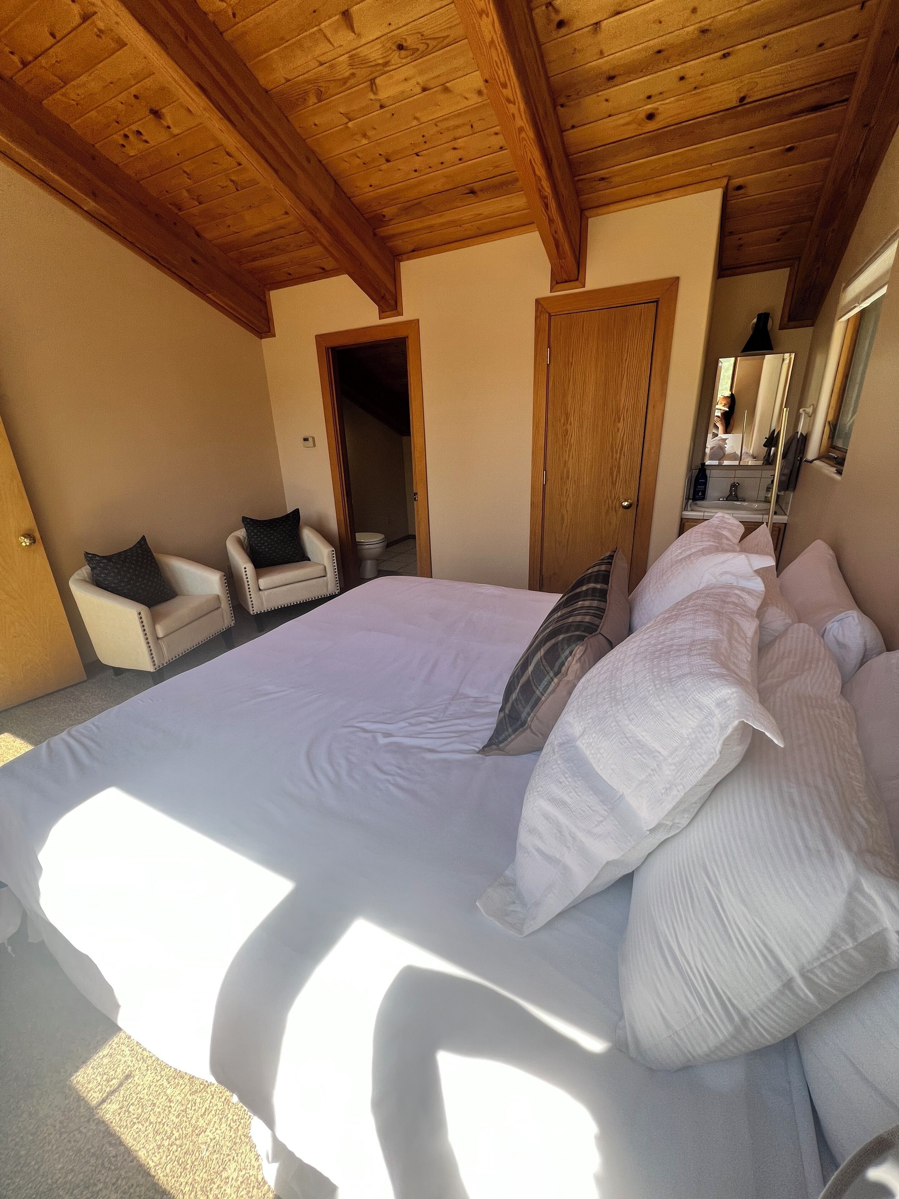 A bedroom with a large bed, a wooden ceiling, a chair, and a door leading to a bathroom.