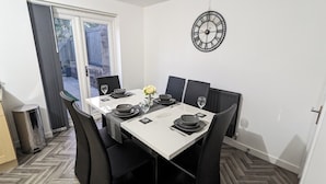 A dining area with a white table set for four, black chairs, a clock on the wall, and a view through a glass door.