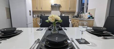 A modern kitchen with wooden cabinets, a built-in oven, and a dining area set with black plates, silverware, and a vase with white flowers.