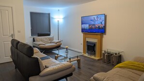 A living room with a sofa, a glass coffee table, a fireplace, and a flat-screen TV mounted above it.