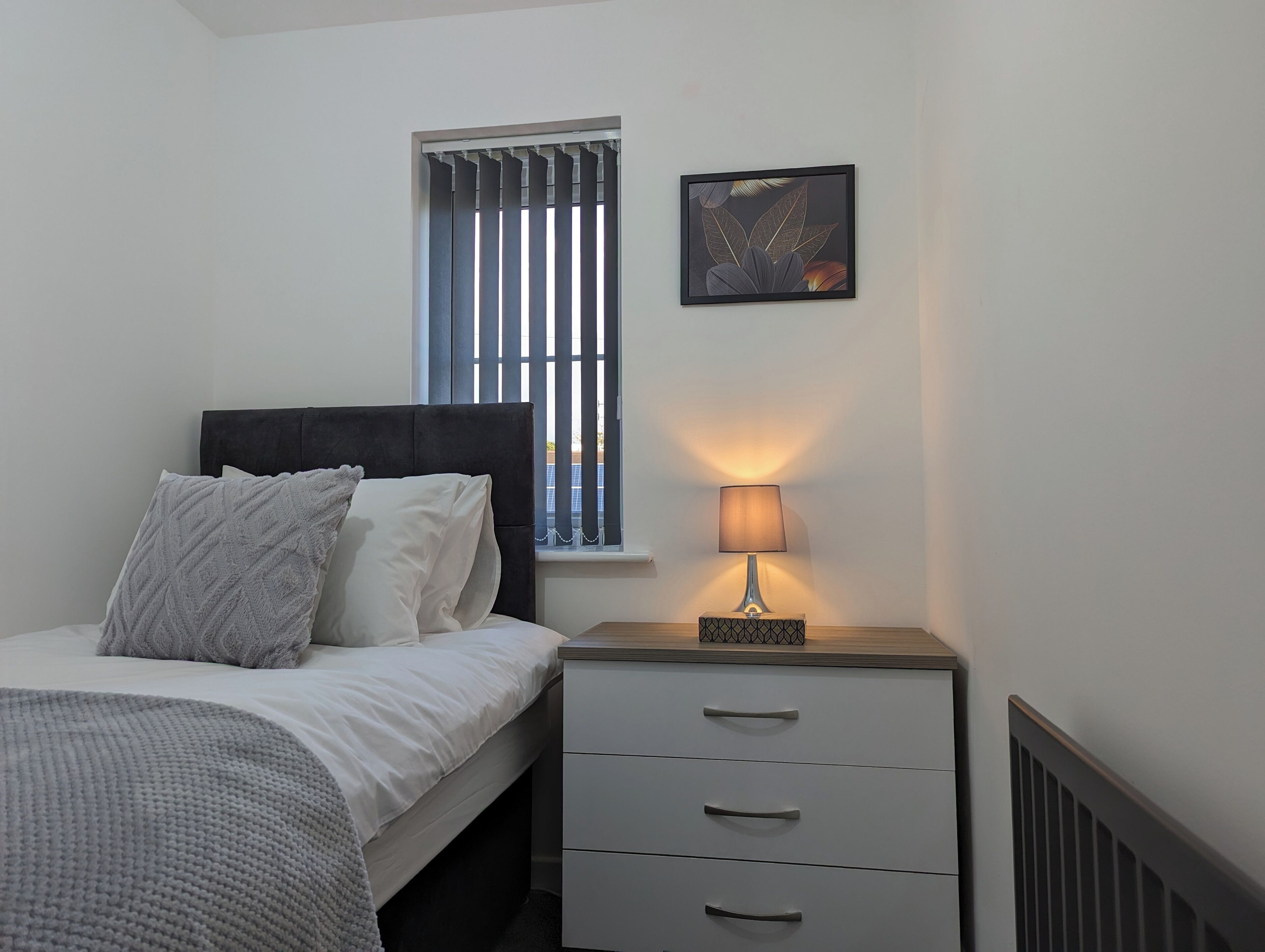 Un dormitorio con cama, mesita de noche con lámpara, ventana con persianas y un cuadro enmarcado en la pared.