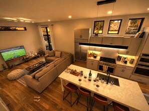 A modern kitchen with a central island, a dining area with bar stools, a living room with a sofa and a TV displaying a sports game, and a large window with curtains.