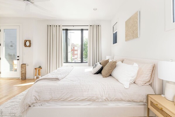 A spacious bedroom with a large bed, a bedside table with a lamp, a small stool, and a view of the outside through a large window.