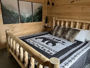 A wooden cabin bedroom with a bed featuring a quilt with a bear design, wooden headboard, and a view of a forest through the window.