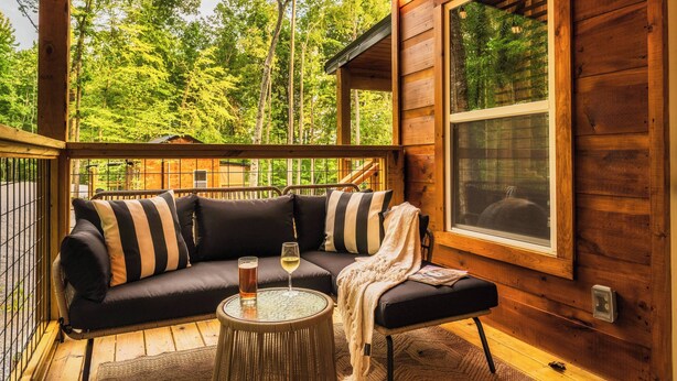 Eine Holzhütte mit einer schwarzen Couch, gestreifteren Kissen, einem Glastisch mit einem Weinglas und Blick auf einen Wald durch ein großes Fenster.