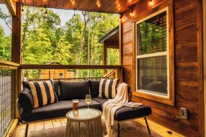 A wooden cabin with a black sofa, striped pillows, a glass table with a glass of wine, and a view of a forest through a large window. - ☆NEW Honeymoon Cabin☆ Hot Tub ◇ Fire Pit ◇ Fireplace ◇ King Bed (Sevierville)