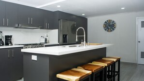 A modern kitchen with dark cabinets, a white countertop, a stainless steel sink, and wooden stools. - CASA TOTALMENTE RENOVADA CON TODA LAS COMODIDADES Y CARACTERISTICAS DE ALTA GAMA (Cali)