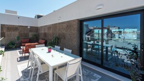 Un patio en la azotea con una mesa y sillas blancas, una puerta de cristal que da a un balcón y plantas en macetas.