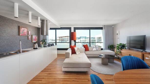 Una sala moderna con sofá, mesa de centro y televisión. Cuenta con una zona de cocina con lavabo y armarios. La habitación tiene ventanas amplias y vista a la ciudad.
