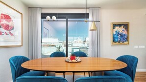 Comedor con una mesa redonda de madera, sillas tapizadas en azul, un centro de mesa rojo y vistas a la ciudad a través de amplias ventanas.