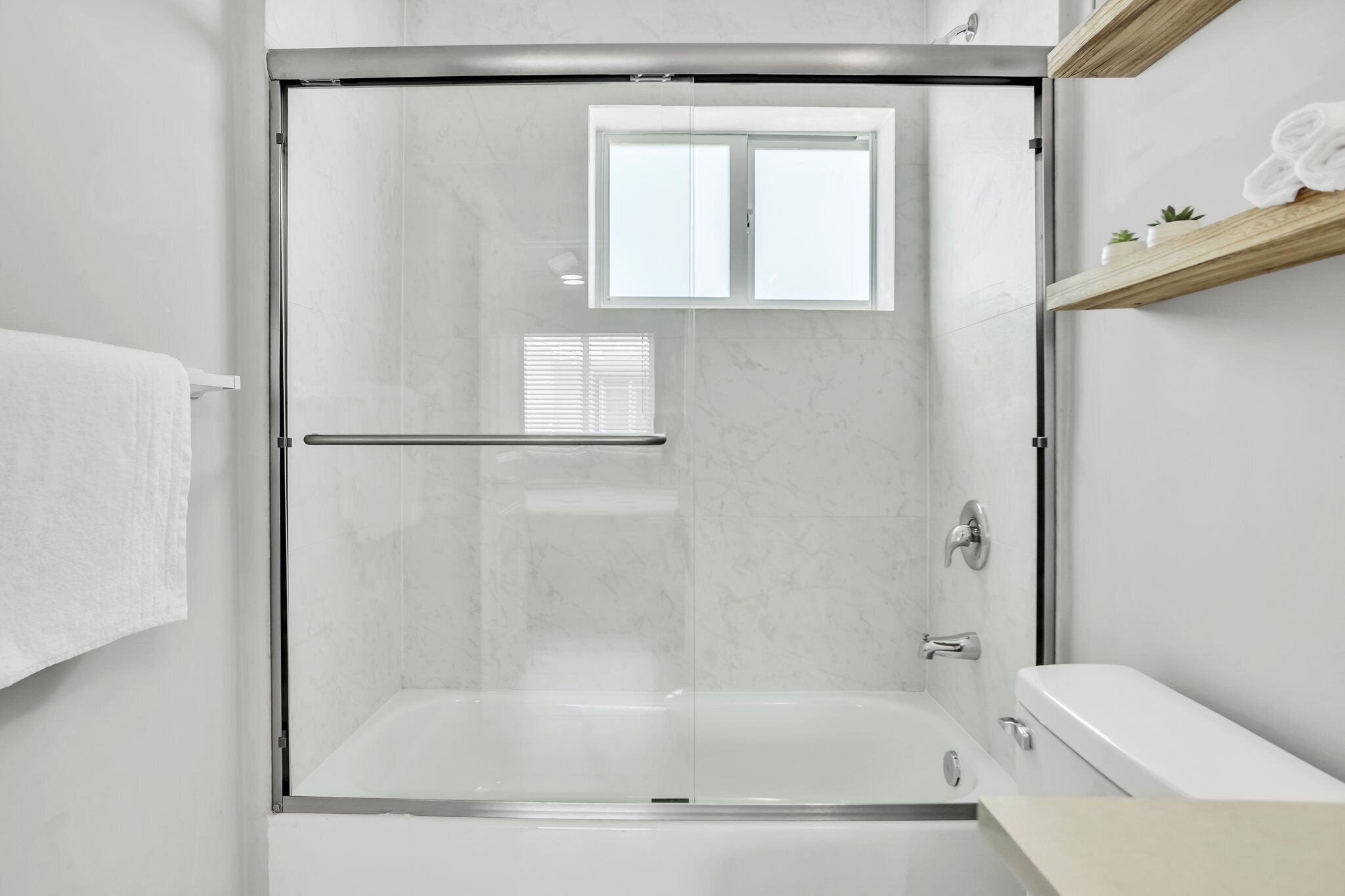 A modern bathroom with a glass-enclosed shower, a white bathtub, and a wooden shelf with towels and plants.