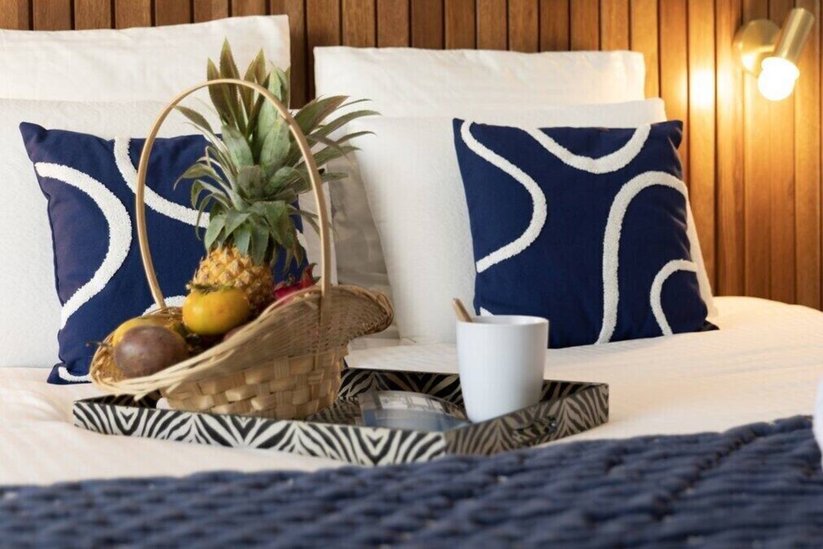 A neatly made bed with blue and white patterned pillows, a woven basket with fruits, a white mug, and a rolled white towel on a dark blue bedspread.