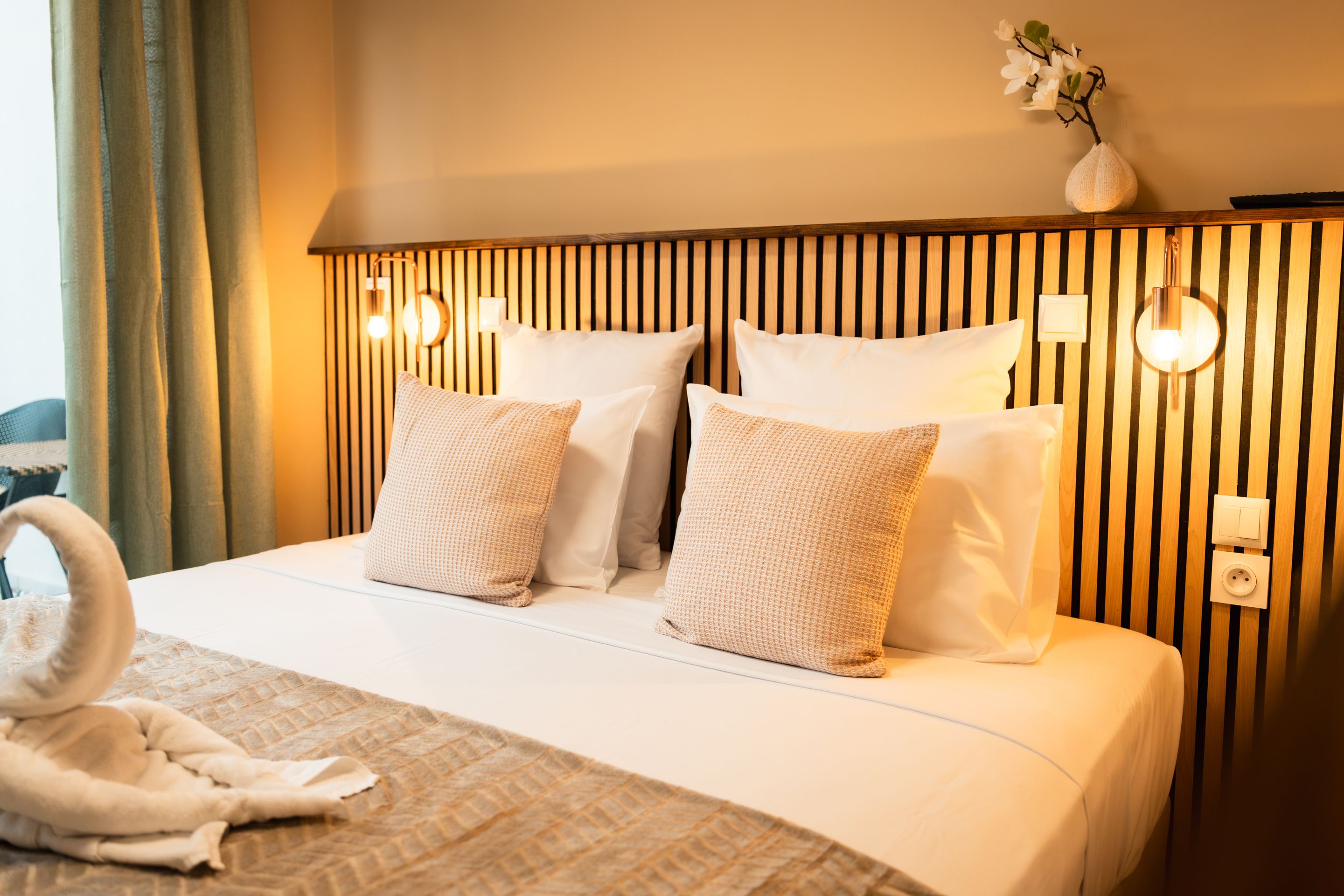 A hotel room with a bed, pillows, a bedside table, a vase with flowers, and a towel on the bed.