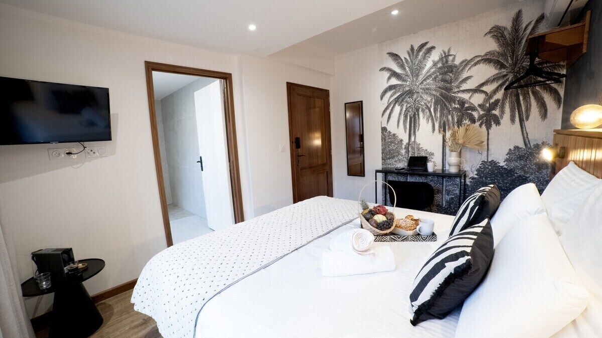 A hotel room with a bed, a TV mounted on the wall, a door, a wall mural, and a small table with a fruit bowl and a cup.