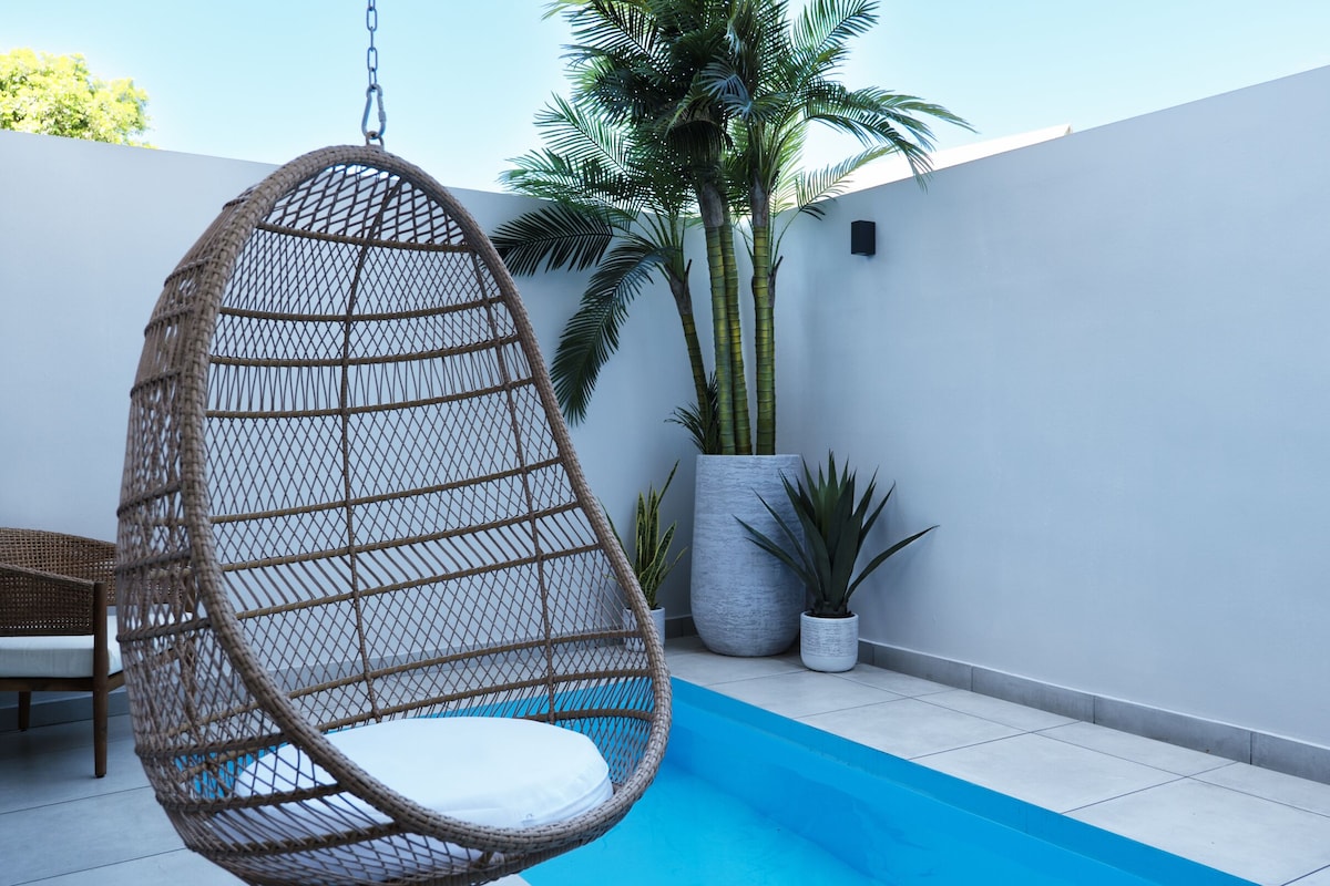 Un fauteuil suspendu près d’une piscine, entouré de plantes en pot et adossé à un mur blanc.