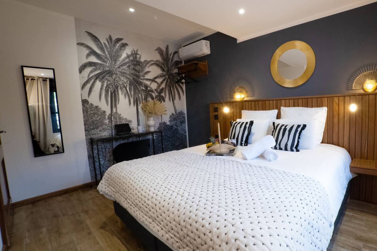 A bedroom with a bed, a wall-mounted mirror, a wall-mounted air conditioner, a decorative wall mural, and a bedside table with a lamp.