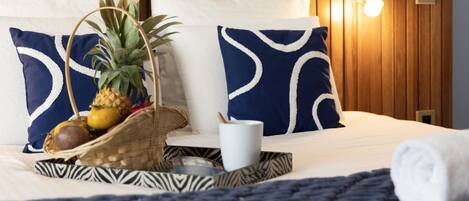 Una cama bien hecha con almohadas de un patrón azul y blanco, una cesta tejida con frutas y una taza blanca sobre la cama.
