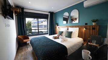 A modern hotel room with a large bed, a desk with a laptop, a chair, a window with curtains, and a view of buildings outside.