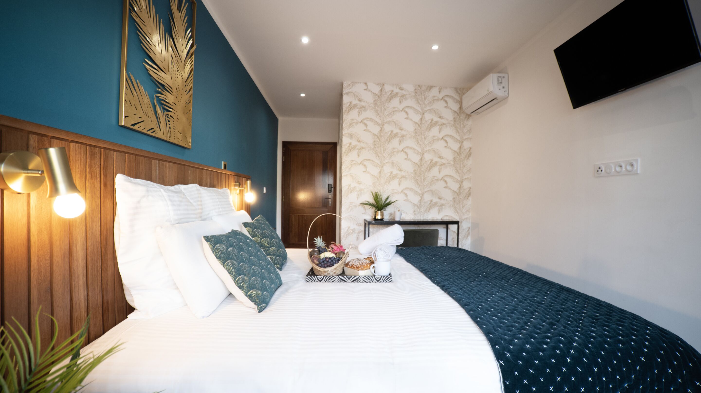 A hotel room with a bed, bedside lamp, wall-mounted TV, and a small table with a plant.