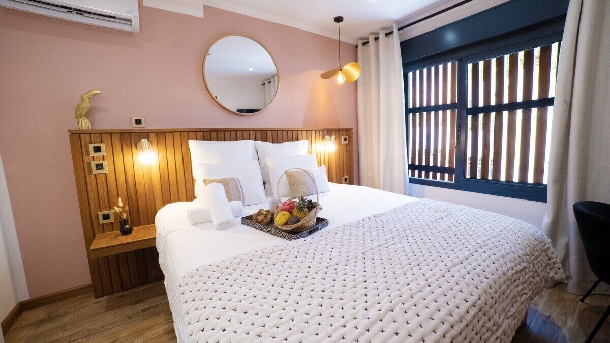 A hotel room with a large bed, a wooden headboard, a round mirror, and a window with blinds.