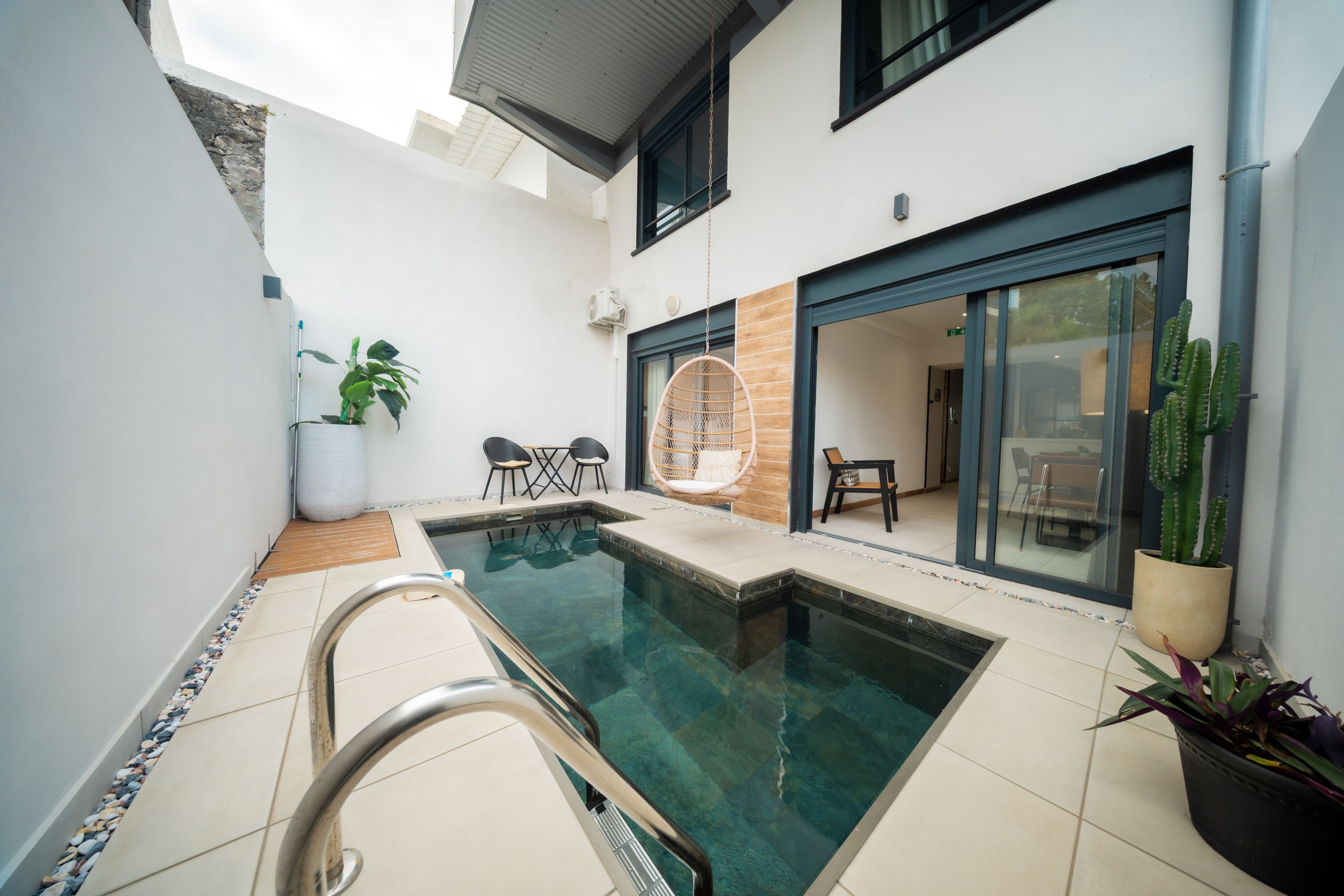 Un área de piscina moderna al aire libre con una piscina cristalina, un espacio de descanso con sillones y una planta en maceta.