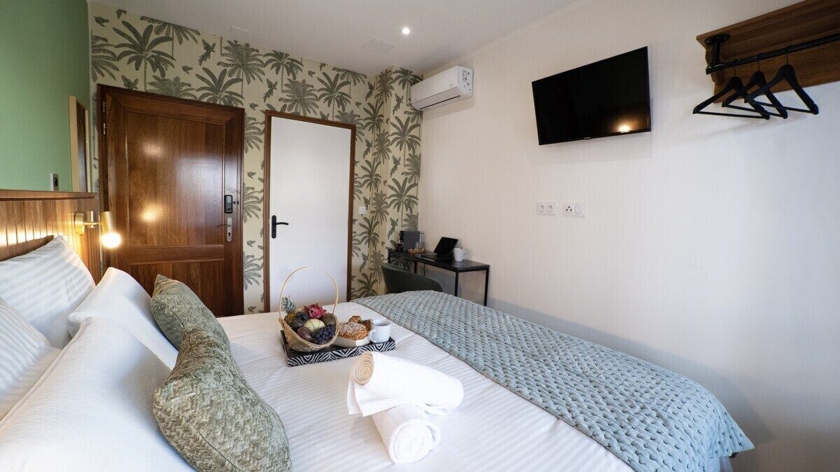 A hotel room with a bed, a small table with a basket of fruits, a TV mounted on the wall, and a coat rack.