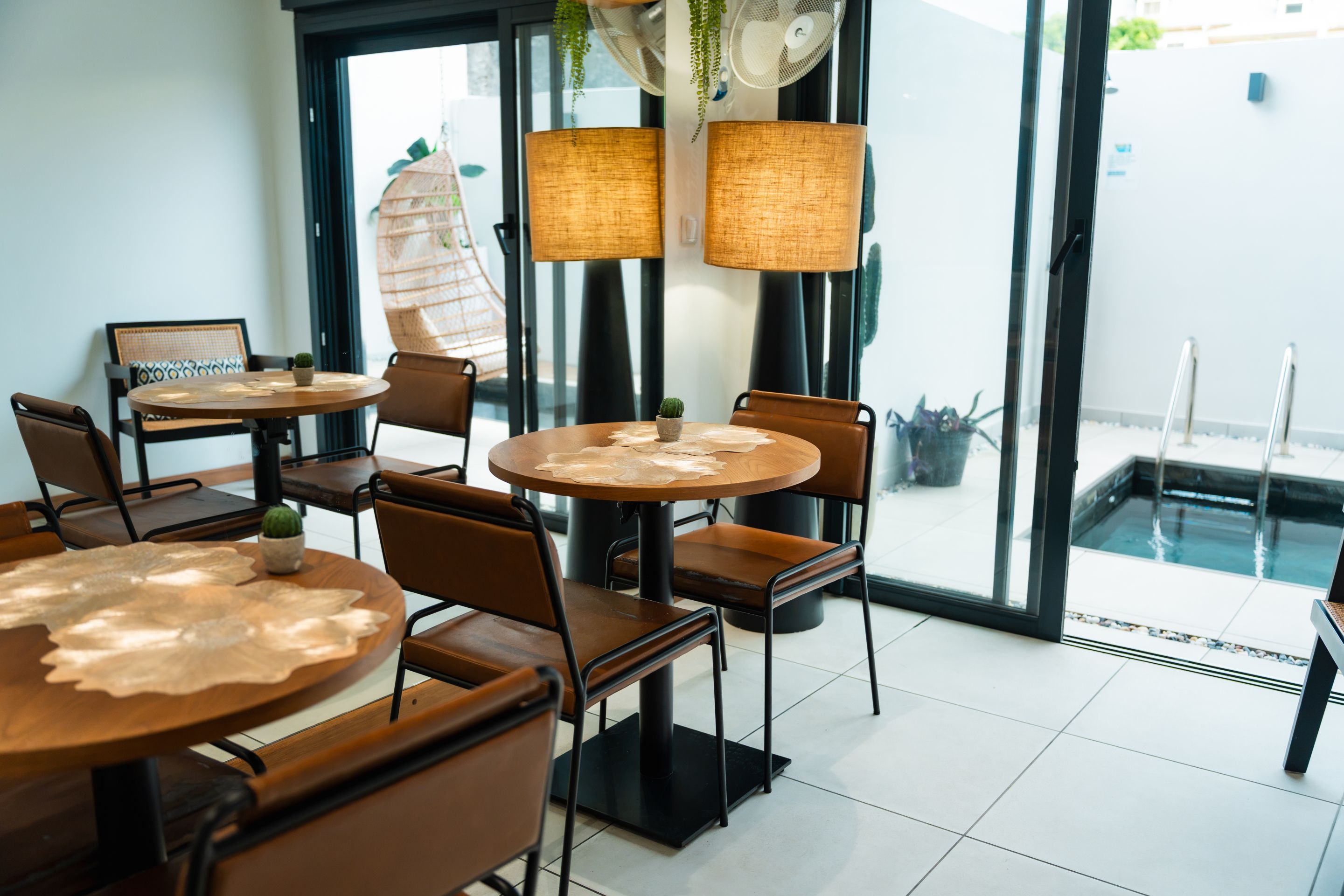 A modern indoor seating area with round tables and chairs, large windows, and a view of a pool.