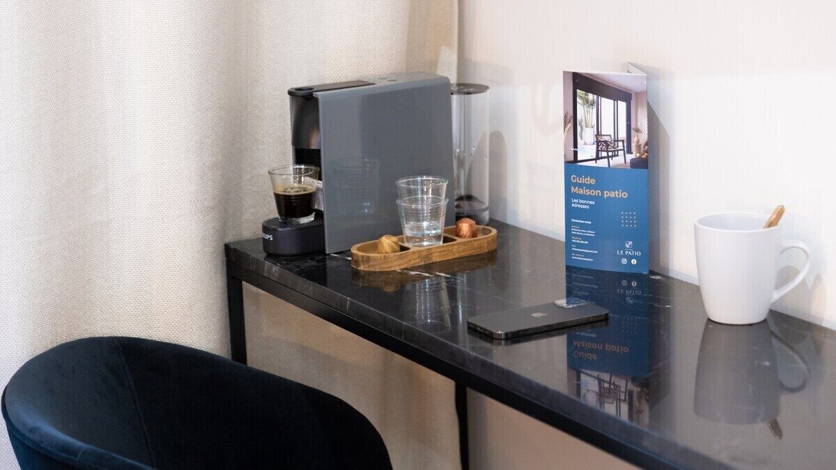 A hotel room with a black table, a coffee machine, a glass of water, a tray with a wooden bowl, a white mug, and a guidebook titled "Guide Maison patio".