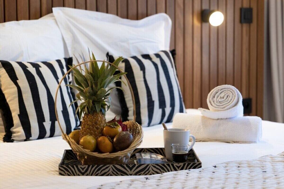 A neatly made bed with a white and black striped pillow, a rolled towel, a fruit bowl with pineapple and oranges, and a tray with a cup and spoon.