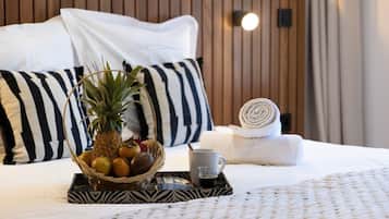 Un lit bien fait, orné d’un oreiller à rayures noires et blanches, une serviette enroulée, un panier de fruits contenant de l’ananas et des oranges, et un plateau avec une tasse et une cuillère.